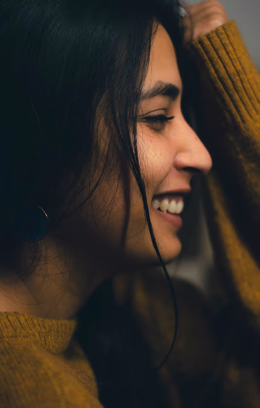 Woman smiling in profile, dark hair and mustard yellow sweater.
